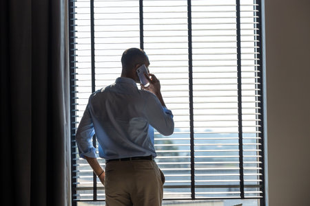 Businessman talking confidently on his mobile phone while standing near large windows in officeの写真素材
