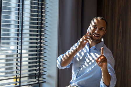 Man wearing shirt posing near window in office and talking on mobile phoneの写真素材