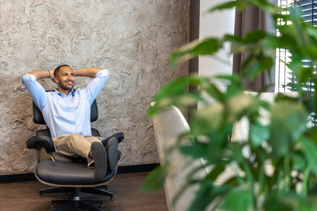 Smiling businessman sitting in chair with raised arms in officeの写真素材
