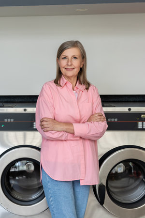 Confident mature woman in pink tshirt standing near washing machines in the laundryの写真素材
