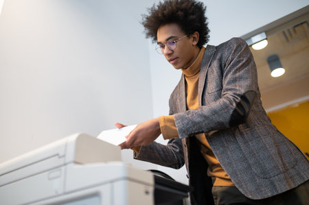 Curly-haired young businessman standing near copier and making documents copies