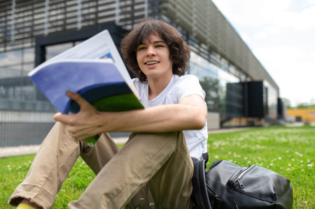 Young male student in casual clothes sitting on grass on street carrying books preparing for school