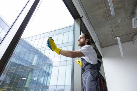 Young service man cleaning windows in the office and looking busyの写真素材