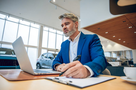 Businessman in suit sitting in automobile dealership office using laptopの写真素材