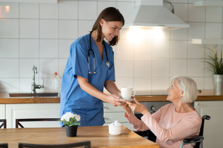 Female nurse helping elderly patient by giving tea offering care and assistanceの写真素材