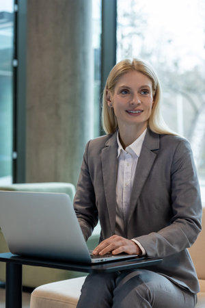 Stylish businesswoman sitting in sofa working with laptop in modern hallの写真素材