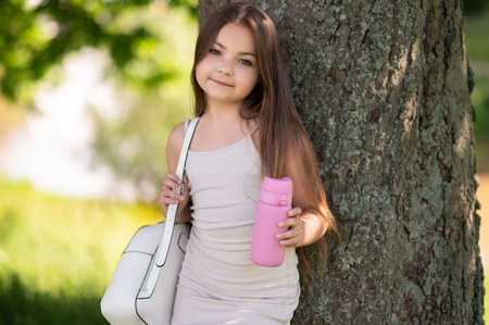 Cute kid standing under tree wearing backpack holding water bottle resting in summer parkの写真素材