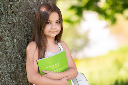 Cute child standing outside in park holding novel book resting in summer parkの写真素材