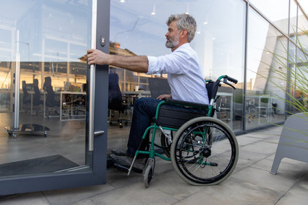 Gray-haired bearded mature man on a wheelchair opening the door to the officeの写真素材
