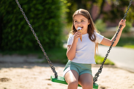 Happy little girl sitting on swing in playground eating sweet ice creamの写真素材