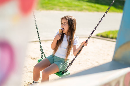 Happy little girl sitting on swing in playground eating sweet ice creamの写真素材