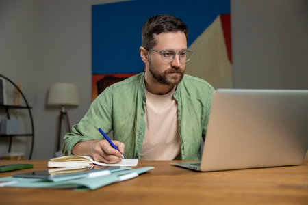 Bearded man reviewing notes on desk while working online with laptop and papersの写真素材
