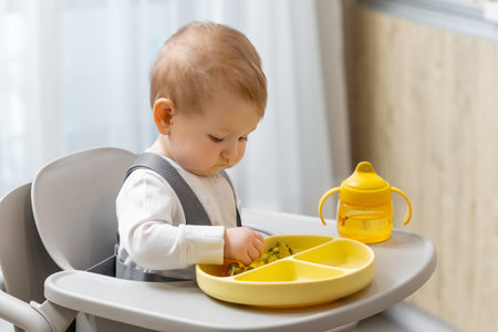 Cute baby boy sitting on his chair and eating from his yellow plateの写真素材