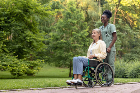 African american nurse rolling a wheelchair of female patient in the parkの写真素材