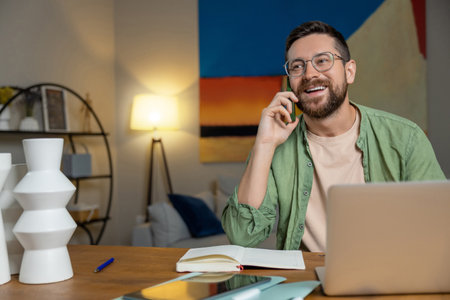 Smiling man talking on phone while working on laptop in modern home officeの写真素材