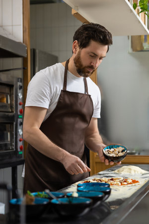 Man works pizza in kitchen adding ingredients carefully preparing foodの写真素材