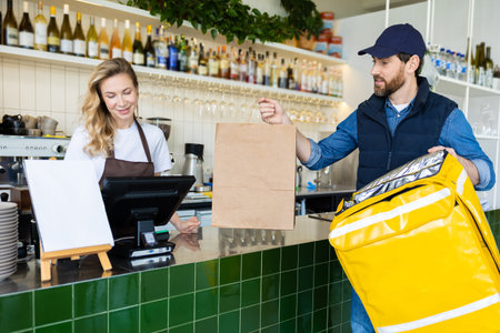 Worker at cafe counter handing order to delivery person in uniform with paper bag