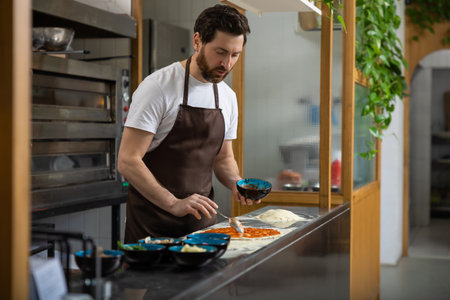 Male cooker working in the pizzeria kitchen and putting topping to the pizza