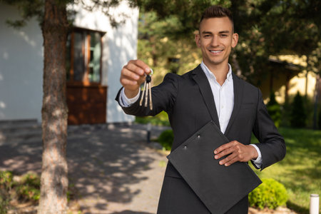 Real estate agent offering house keys and paperwork on clipboard in front of residential propertyの写真素材
