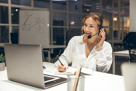 Office worker managing tasks on laptop with headset in bright roomの写真素材