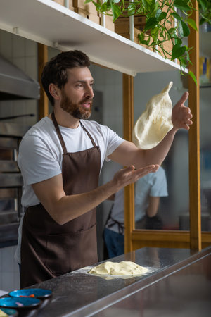 Bearded man baker wearing white T-snirt and brown apron working with dough for pizza