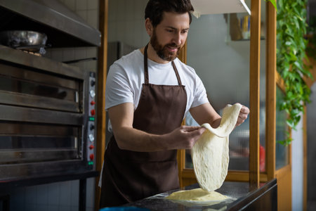 Bearded man wearing a white t-shirt and brown apron tosses pizza dough in a bakery kitchenの写真素材