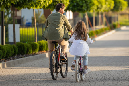 Back view of family bike ride with father and daughter on park path near homeの写真素材