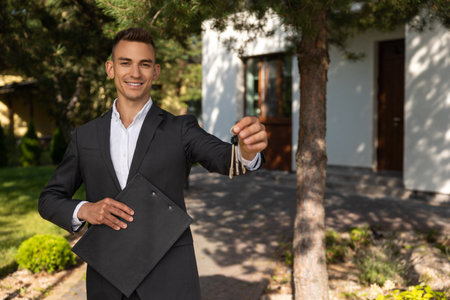 Real estate agent holding keys and clipboard in front of modern house for propertyの写真素材