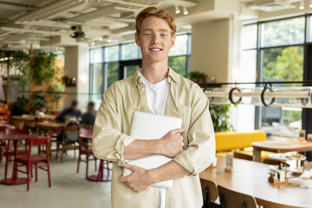 Positive young man with a laptop in hands standing in a cafeの写真素材