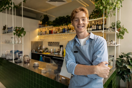 Cheerful young cafÃ© worker with crossed arms leaning against counter in stylish interior