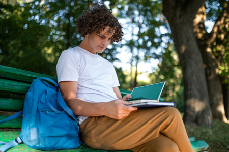 Curly-haired teenager sitting on the bench in the park and reading a book