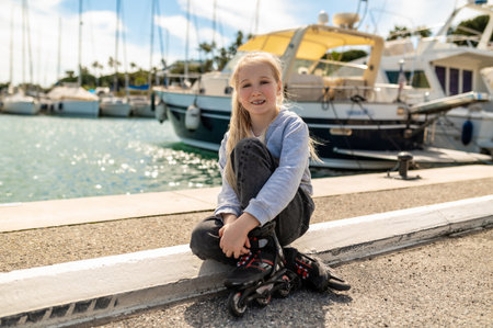 Blonde girl in inline skates sitting cross legged on dockside near moored sailboats and calm seaの写真素材