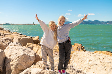 Happy children sisters posing on rocks near sea on sunny rocky coastline day with raised armsの写真素材