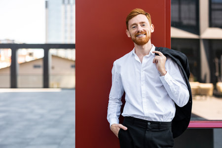 Ginger bearded man in white shirt posing outside by red building wallの写真素材
