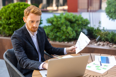 Ginger-haired man in suit reading reports while using laptop outdoorの写真素材