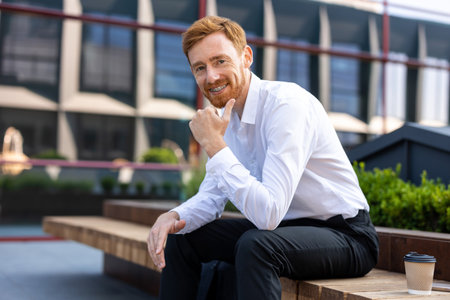Handsome bearded man with ginger hair dressed in white shirt resting on bench in open airの写真素材