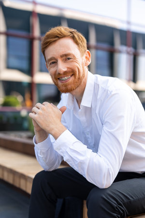 Confident ginger man with beard wearing white shirt sitting outdoors on city benchの写真素材