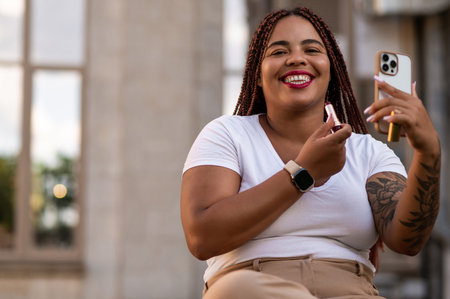 Confident plus size woman applying lipstick while looking at smartphone sittingの写真素材