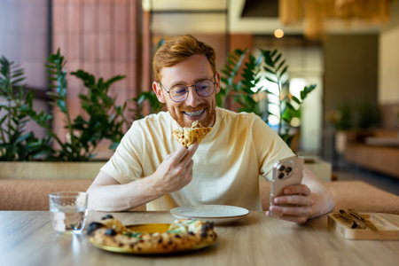 Ginger bearded young adult in glasses holding phone and enjoying pizza