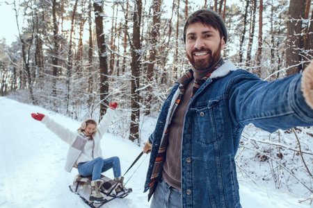 Bearded young man pulling sled with his wife sitting on it