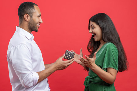 Cheerful couple celebrating birthday together on red background man presenting gift box to womanの写真素材