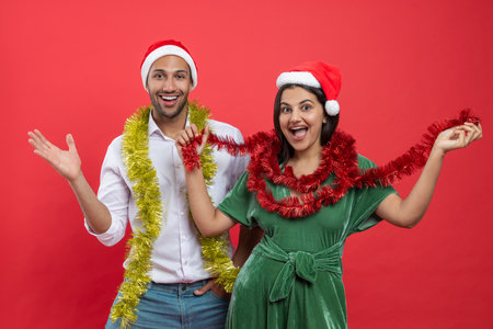 Cheerful couple in festive Santa hats and tinsel over necks celebrating Christmas and New Year