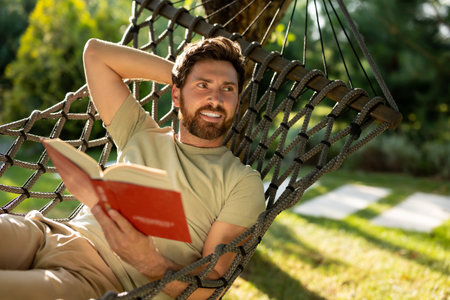 Caucasian bearded young man lying in hammock with a book in handsの写真素材