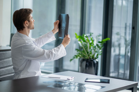 Male doctor sitting at desk looking at x-ray in modern clinic with windowの写真素材