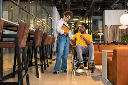 Woman holding folders and man in wheelchair discussing project in coworking space