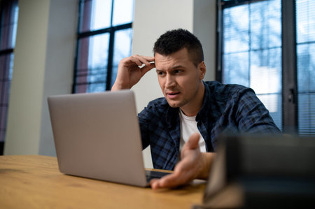 Concentrated man touching forehead while working on laptop at wooden table in office