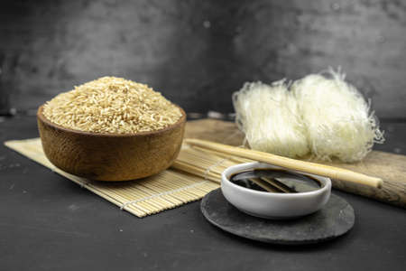 brown rice in a wooden bowl, asian kitchen background with bamboo mat, chopsticks, soy sauce, pickled ginger and glass rice noodles on black backgroundの写真素材