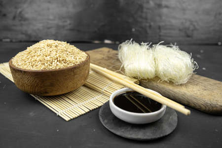 brown rice in a wooden bowl, asian kitchen background with bamboo mat, chopsticks, soy sauce, pickled ginger and glass rice noodles on black backgroundの写真素材