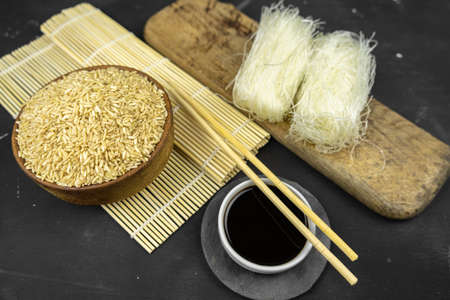 brown rice in a wooden bowl, asian kitchen background with bamboo mat, chopsticks, soy sauce, pickled ginger and glass rice noodles on black backgroundの写真素材
