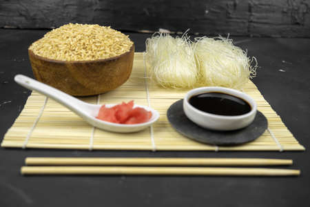 brown rice in a wooden bowl, asian kitchen background with bamboo mat, chopsticks, soy sauce, pickled ginger and glass rice noodles on black backgroundの写真素材